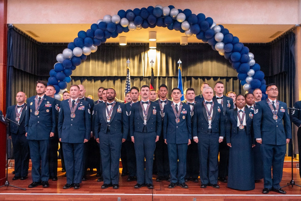 A formal military graduation or awards ceremony featuring approximately 25-30 uniformed personnel standing on a stage. The individuals are wearing dark blue dress uniforms with medals and ribbons, posed beneath a decorative arch of blue and silver balloons. Multiple flags are visible in the background, including the American flag and what appears to be military unit flags.