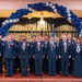 A formal military graduation or awards ceremony featuring approximately 25-30 uniformed personnel standing on a stage. The individuals are wearing dark blue dress uniforms with medals and ribbons, posed beneath a decorative arch of blue and silver balloons. Multiple flags are visible in the background, including the American flag and what appears to be military unit flags.
