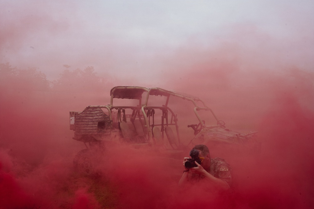 A photographer in camouflage uniform is capturing images amid dense red smoke, likely from a smoke grenade or flare used during a military exercise or training scenario. Behind the photographer is a multi-seat military utility vehicle (UTV/MRZR-type tactical vehicle) partially obscured by the red smoke. The scene suggests a military documentation or training exercise environment.
