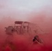 A photographer in camouflage uniform is capturing images amid dense red smoke, likely from a smoke grenade or flare used during a military exercise or training scenario. Behind the photographer is a multi-seat military utility vehicle (UTV/MRZR-type tactical vehicle) partially obscured by the red smoke. The scene suggests a military documentation or training exercise environment.