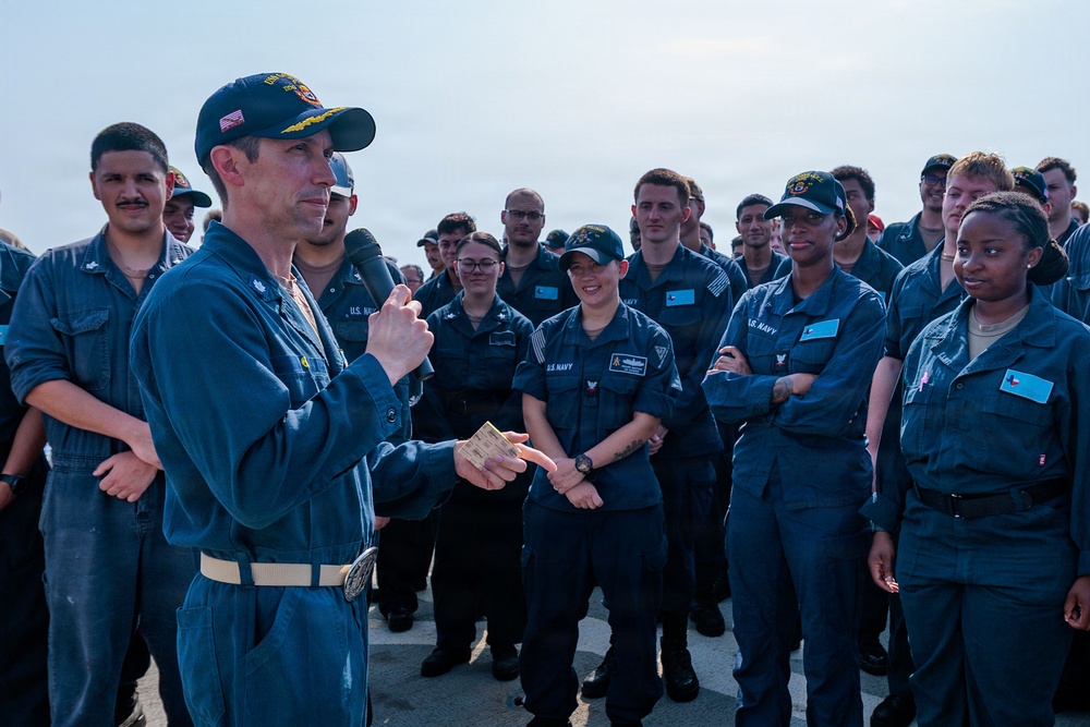 A senior U.S. Navy officer, wearing a command ball cap with aviator wings insignia and holding a microphone, addresses a large group of enlisted sailors in blue coverall uniforms. The gathering appears to be an all-hands or crew briefing taking place on what looks like a flight deck or outdoor deck of a naval vessel. The sailors are attentive and some are visibly smiling, suggesting a positive or motivational address.