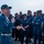 A senior U.S. Navy officer, wearing a command ball cap with aviator wings insignia and holding a microphone, addresses a large group of enlisted sailors in blue coverall uniforms. The gathering appears to be an all-hands or crew briefing taking place on what looks like a flight deck or outdoor deck of a naval vessel. The sailors are attentive and some are visibly smiling, suggesting a positive or motivational address.
