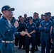 A senior U.S. Navy officer, wearing a command ball cap with aviator wings insignia and holding a microphone, addresses a large group of enlisted sailors in blue coverall uniforms. The gathering appears to be an all-hands or crew briefing taking place on what looks like a flight deck or outdoor deck of a naval vessel. The sailors are attentive and some are visibly smiling, suggesting a positive or motivational address.