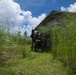 Two armed personnel in military/tactical camouflage gear are positioned near the exterior wall of a weathered building surrounded by tall grass. One individual is standing and holding a rifle upward while the other is in a kneeling position, suggesting a tactical clearing or security operation. The setting appears to be an overgrown, possibly abandoned structure in a tropical or subtropical environment.