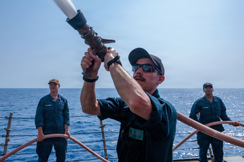 Three U.S. Navy sailors are on the deck of a naval vessel at sea, conducting what appears to be a firefighting drill or water hose exercise. The central figure is actively operating a large fire hose nozzle, directing it upward, while two other sailors observe in the background. The setting is an open ocean environment under clear, sunny skies.