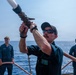 Three U.S. Navy sailors are on the deck of a naval vessel at sea, conducting what appears to be a firefighting drill or water hose exercise. The central figure is actively operating a large fire hose nozzle, directing it upward, while two other sailors observe in the background. The setting is an open ocean environment under clear, sunny skies.