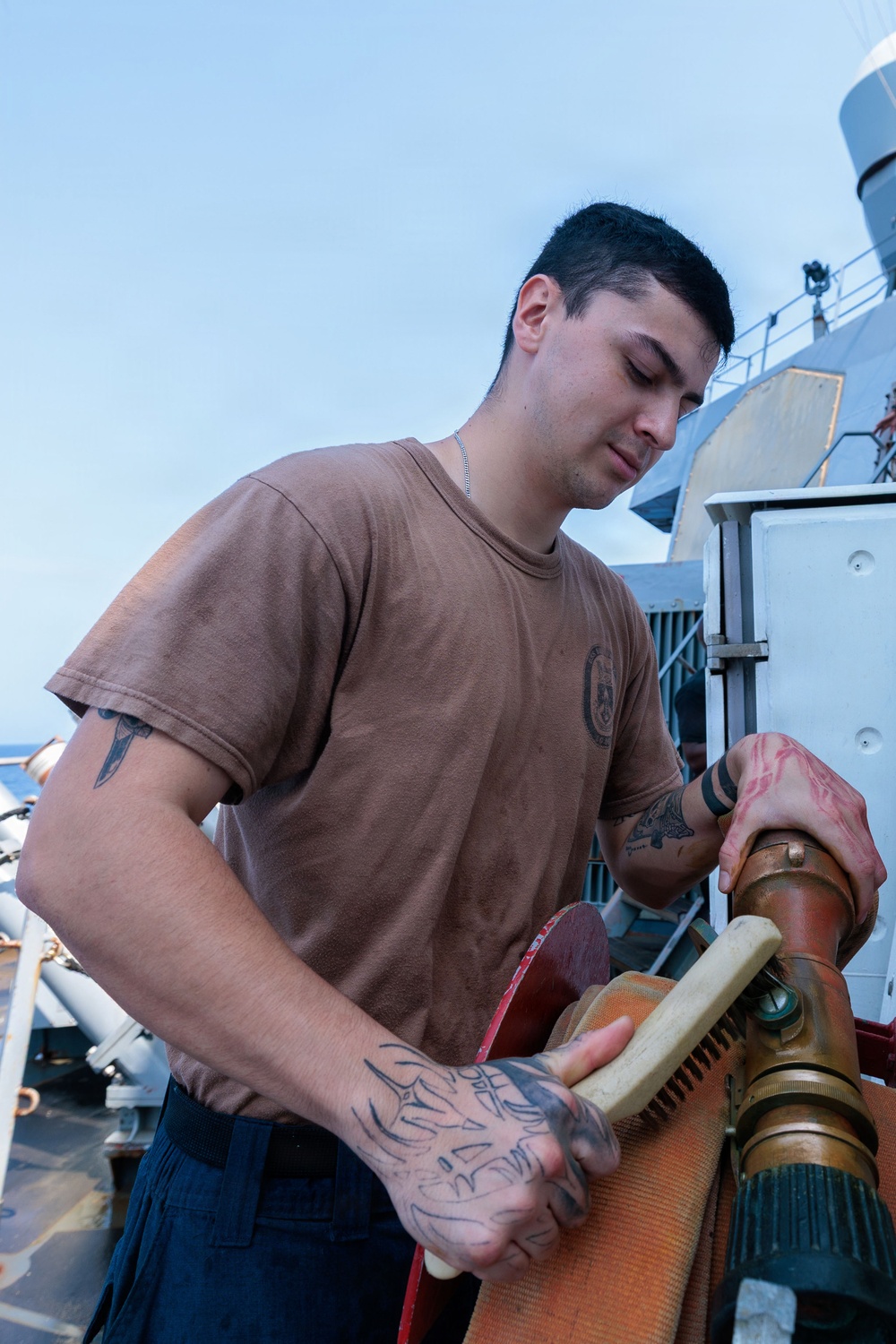 A young male sailor or service member is working on the deck of a naval vessel, handling what appears to be a fire hose or similar equipment connected to a brass fitting. He is wearing a brown military-style t-shirt with a small emblem on the chest and dark navy trousers. The individual has multiple visible tattoos on both hands and arms.