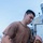 A young male sailor or service member is working on the deck of a naval vessel, handling what appears to be a fire hose or similar equipment connected to a brass fitting. He is wearing a brown military-style t-shirt with a small emblem on the chest and dark navy trousers. The individual has multiple visible tattoos on both hands and arms.