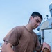 A young male sailor or service member is working on the deck of a naval vessel, handling what appears to be a fire hose or similar equipment connected to a brass fitting. He is wearing a brown military-style t-shirt with a small emblem on the chest and dark navy trousers. The individual has multiple visible tattoos on both hands and arms.