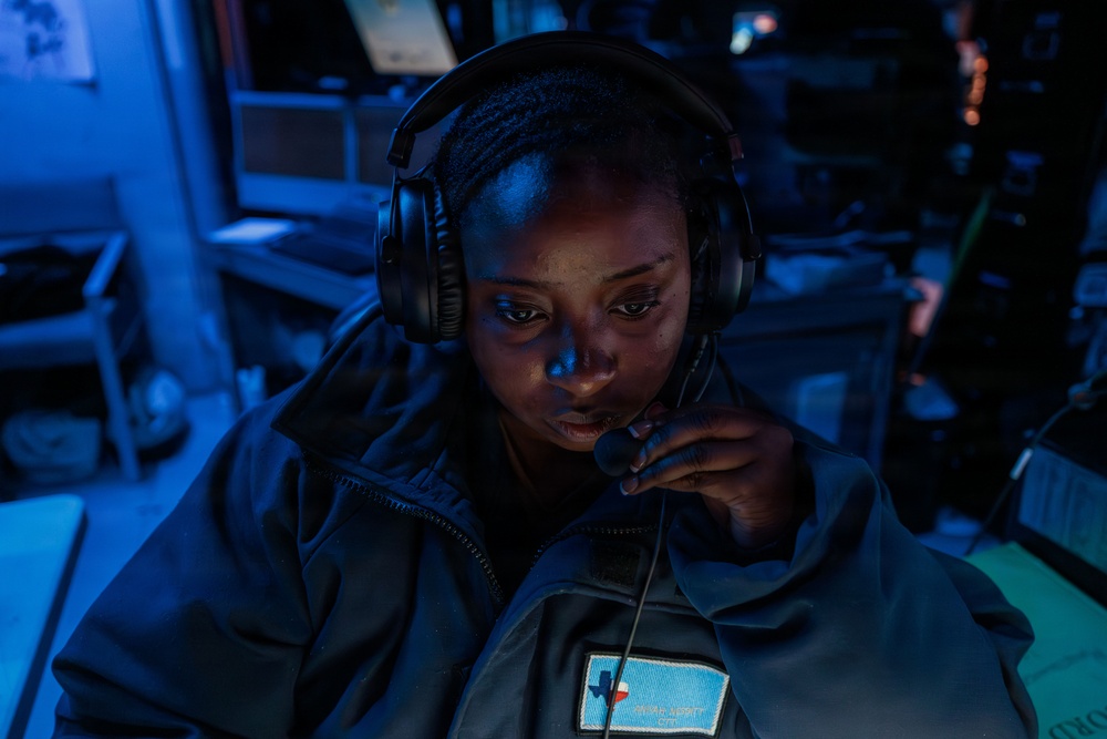 A woman wearing a headset with a boom microphone is seated in a dimly lit operational environment illuminated by blue lighting. She is wearing a dark jacket with a patch featuring the Texas state outline on a light blue background. She appears to be actively communicating or monitoring, holding the microphone close to her mouth.