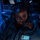 A woman wearing a headset with a boom microphone is seated in a dimly lit operational environment illuminated by blue lighting. She is wearing a dark jacket with a patch featuring the Texas state outline on a light blue background. She appears to be actively communicating or monitoring, holding the microphone close to her mouth.