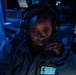 A woman wearing a headset with a boom microphone is seated in a dimly lit operational environment illuminated by blue lighting. She is wearing a dark jacket with a patch featuring the Texas state outline on a light blue background. She appears to be actively communicating or monitoring, holding the microphone close to her mouth.