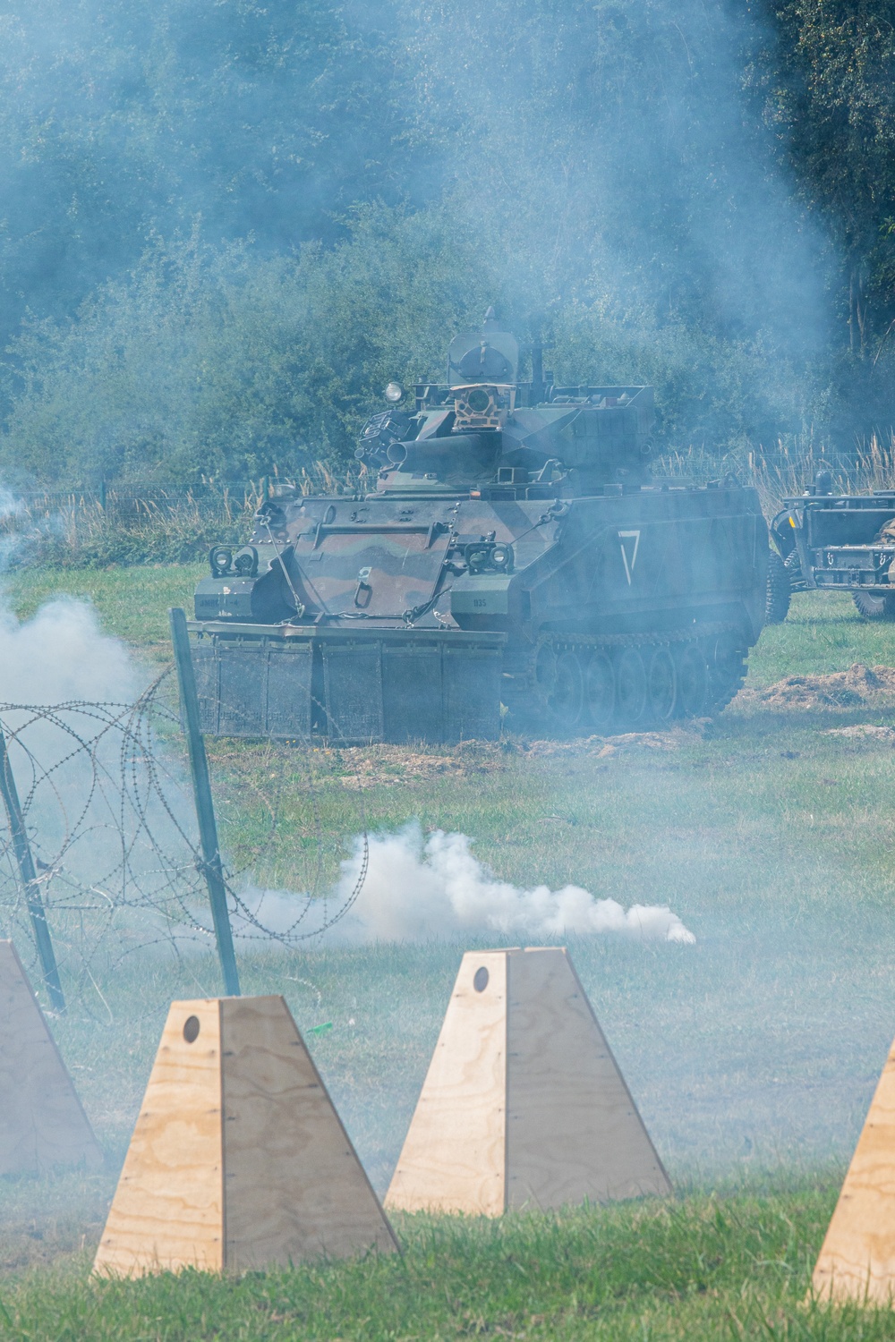 A tracked armored personnel carrier (APC) in camouflage paint advances through a smoke-filled field during what appears to be a military exercise or demonstration. The vehicle is approaching a defensive obstacle line consisting of wooden anti-tank barriers and barbed wire concertina fencing. Smoke grenades or pyrotechnic devices are actively producing white smoke around the scene.