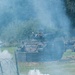 A tracked armored personnel carrier (APC) in camouflage paint advances through a smoke-filled field during what appears to be a military exercise or demonstration. The vehicle is approaching a defensive obstacle line consisting of wooden anti-tank barriers and barbed wire concertina fencing. Smoke grenades or pyrotechnic devices are actively producing white smoke around the scene.