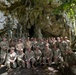 A large group of military personnel in camouflage uniforms are posing for a group photograph in front of a large limestone cave entrance surrounded by tropical jungle vegetation. The group consists of approximately 25-30 individuals arranged in multiple rows, with some kneeling in the front. The setting appears to be a tropical or subtropical environment based on the lush green foliage visible around the cave.