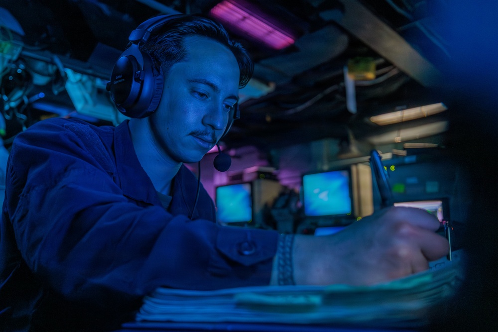 A military or naval personnel member wearing a headset with microphone is working at a dimly lit operations station, illuminated primarily by blue ambient lighting. The individual is focused on documents or equipment in front of them, surrounded by multiple monitor screens in the background. The setting appears to be a combat information center or similar shipboard operations room.