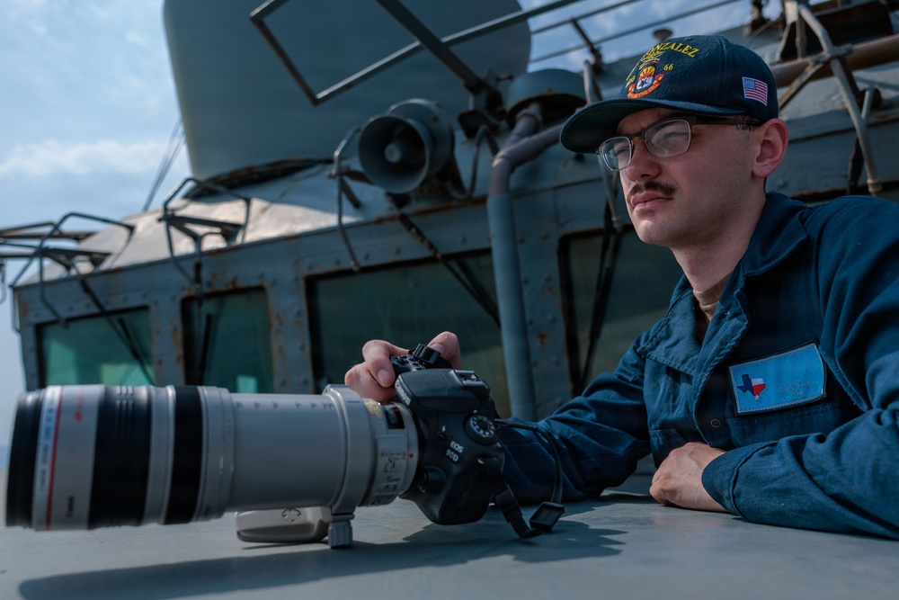 A U.S. Navy sailor is positioned on the deck of a naval vessel, operating a Canon EOS 90D DSLR camera equipped with a large white telephoto lens. The sailor wears a dark blue uniform with a Texas state patch on the chest and a ship's ball cap reading 'GONZALEZ 66' with an American flag patch. The setting is clearly aboard a military ship, with gray naval superstructure and equipment visible in the background.
