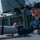 A U.S. Navy sailor is positioned on the deck of a naval vessel, operating a Canon EOS 90D DSLR camera equipped with a large white telephoto lens. The sailor wears a dark blue uniform with a Texas state patch on the chest and a ship's ball cap reading 'GONZALEZ 66' with an American flag patch. The setting is clearly aboard a military ship, with gray naval superstructure and equipment visible in the background.