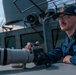 A U.S. Navy sailor is positioned on the deck of a naval vessel, operating a Canon EOS 90D DSLR camera equipped with a large white telephoto lens. The sailor wears a dark blue uniform with a Texas state patch on the chest and a ship's ball cap reading 'GONZALEZ 66' with an American flag patch. The setting is clearly aboard a military ship, with gray naval superstructure and equipment visible in the background.