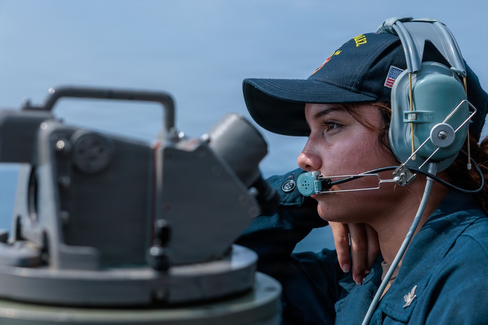 A female U.S. Navy sailor is stationed on the deck of a naval vessel, wearing a dark blue Navy cap with an American flag patch and a gray communications headset with boom microphone. She appears to be standing watch or performing navigational duties near what appears to be a gyrocompass or alidade sighting instrument. The background shows open water and a hazy sky, consistent with an at-sea operational environment.
