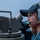 A female U.S. Navy sailor is stationed on the deck of a naval vessel, wearing a dark blue Navy cap with an American flag patch and a gray communications headset with boom microphone. She appears to be standing watch or performing navigational duties near what appears to be a gyrocompass or alidade sighting instrument. The background shows open water and a hazy sky, consistent with an at-sea operational environment.