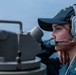 A female U.S. Navy sailor is stationed on the deck of a naval vessel, wearing a dark blue Navy cap with an American flag patch and a gray communications headset with boom microphone. She appears to be standing watch or performing navigational duties near what appears to be a gyrocompass or alidade sighting instrument. The background shows open water and a hazy sky, consistent with an at-sea operational environment.