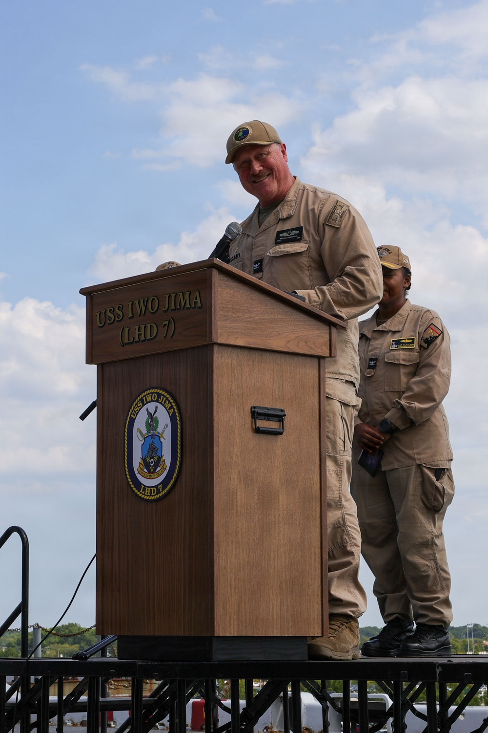 A military officer in tan/khaki uniform is speaking at a podium bearing the USS Iwo Jima (LHD 7) insignia and ship's seal. A second uniformed service member stands behind and to the right of the speaker. The event appears to be an official naval ceremony or address conducted outdoors under a partly cloudy sky.