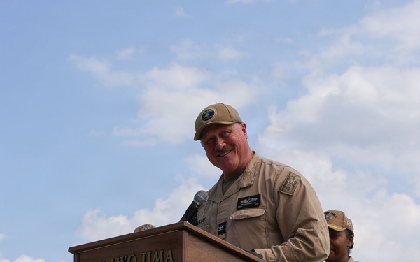 A military officer in tan/khaki uniform is speaking at a podium bearing the USS Iwo Jima (LHD 7) insignia and ship's seal. A second uniformed service member stands behind and to the right of the speaker. The event appears to be an official naval ceremony or address conducted outdoors under a partly cloudy sky.