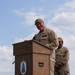 A military officer in tan/khaki uniform is speaking at a podium bearing the USS Iwo Jima (LHD 7) insignia and ship's seal. A second uniformed service member stands behind and to the right of the speaker. The event appears to be an official naval ceremony or address conducted outdoors under a partly cloudy sky.