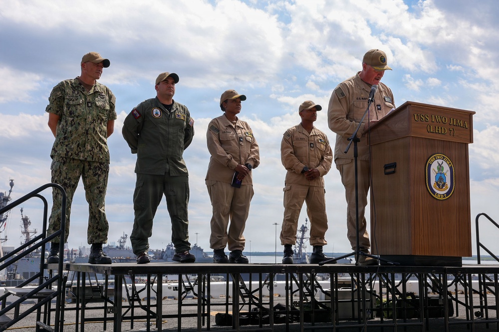 A military ceremony is taking place aboard the USS Iwo Jima (LHD 7), an amphibious assault ship. Five military personnel in various uniforms are on a raised platform, with one individual speaking at a podium bearing the ship's name and insignia. Naval vessels and a harbor are visible in the background.