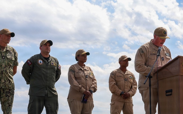 A military ceremony is taking place aboard the USS Iwo Jima (LHD 7), an amphibious assault ship. Five military personnel in various uniforms are on a raised platform, with one individual speaking at a podium bearing the ship's name and insignia. Naval vessels and a harbor are visible in the background.