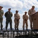A military ceremony is taking place aboard the USS Iwo Jima (LHD 7), an amphibious assault ship. Five military personnel in various uniforms are on a raised platform, with one individual speaking at a podium bearing the ship's name and insignia. Naval vessels and a harbor are visible in the background.