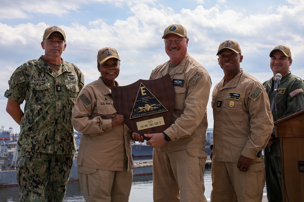 Five U.S. Navy personnel pose together holding a wooden shield-shaped plaque identified as the 'Battle Effectiveness Award' from Commander Naval Surface Forces Atlantic, awarded to USS Iwo Jima (LHD-7) in 2025. The group includes personnel in various uniforms — one in camouflage NWU Type III, three in tan/khaki working uniforms, and one in a flight suit — photographed outdoors at a naval installation with ships visible in the background.