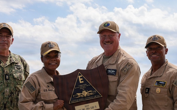 Five U.S. Navy personnel pose together holding a wooden shield-shaped plaque identified as the 'Battle Effectiveness Award' from Commander Naval Surface Forces Atlantic, awarded to USS Iwo Jima (LHD-7) in 2025. The group includes personnel in various uniforms — one in camouflage NWU Type III, three in tan/khaki working uniforms, and one in a flight suit — photographed outdoors at a naval installation with ships visible in the background.