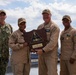 Five U.S. Navy personnel pose together holding a wooden shield-shaped plaque identified as the 'Battle Effectiveness Award' from Commander Naval Surface Forces Atlantic, awarded to USS Iwo Jima (LHD-7) in 2025. The group includes personnel in various uniforms — one in camouflage NWU Type III, three in tan/khaki working uniforms, and one in a flight suit — photographed outdoors at a naval installation with ships visible in the background.