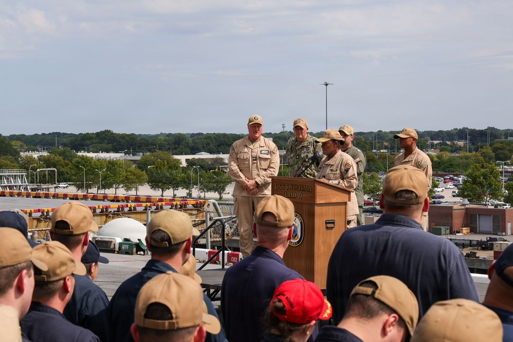 A military ceremony is taking place on the deck of the USS Iwo Jima (LHD-7), an amphibious assault ship. Several uniformed personnel are gathered around a wooden podium bearing the ship's name and insignia, with one individual appearing to speak or present. An audience of sailors and military personnel in tan and navy uniforms observe from the foreground.