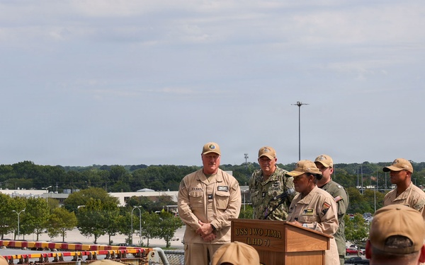 A military ceremony is taking place on the deck of the USS Iwo Jima (LHD-7), an amphibious assault ship. Several uniformed personnel are gathered around a wooden podium bearing the ship's name and insignia, with one individual appearing to speak or present. An audience of sailors and military personnel in tan and navy uniforms observe from the foreground.