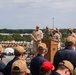 A military ceremony is taking place on the deck of the USS Iwo Jima (LHD-7), an amphibious assault ship. Several uniformed personnel are gathered around a wooden podium bearing the ship's name and insignia, with one individual appearing to speak or present. An audience of sailors and military personnel in tan and navy uniforms observe from the foreground.