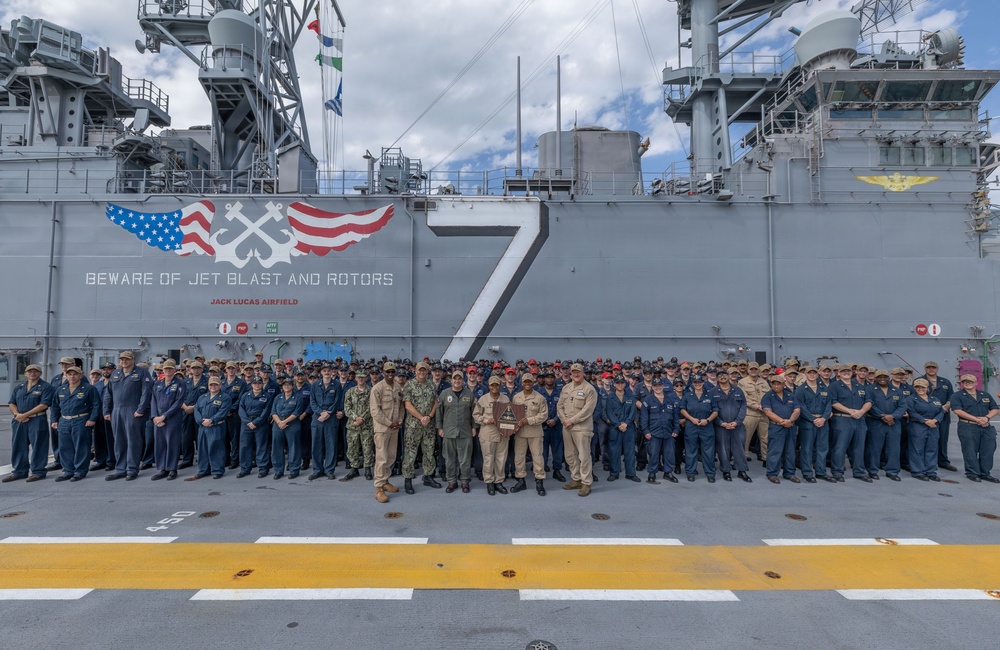 A large group of U.S. Navy personnel are assembled on the flight deck of a naval vessel, identified as 'Jack Lucas Airfield' with hull number 7, for what appears to be an award or recognition ceremony. The ship's island superstructure is visible in the background, displaying the warning 'BEWARE OF JET BLAST AND ROTORS' along with a decorative emblem featuring American flags and crossed anchors. Several individuals in the center of the group are holding what appears to be a trophy or award plaque.