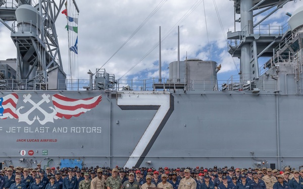 A large group of U.S. Navy personnel are assembled on the flight deck of a naval vessel, identified as 'Jack Lucas Airfield' with hull number 7, for what appears to be an award or recognition ceremony. The ship's island superstructure is visible in the background, displaying the warning 'BEWARE OF JET BLAST AND ROTORS' along with a decorative emblem featuring American flags and crossed anchors. Several individuals in the center of the group are holding what appears to be a trophy or award plaque.