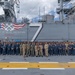 A large group of U.S. Navy personnel are assembled on the flight deck of a naval vessel, identified as 'Jack Lucas Airfield' with hull number 7, for what appears to be an award or recognition ceremony. The ship's island superstructure is visible in the background, displaying the warning 'BEWARE OF JET BLAST AND ROTORS' along with a decorative emblem featuring American flags and crossed anchors. Several individuals in the center of the group are holding what appears to be a trophy or award plaque.