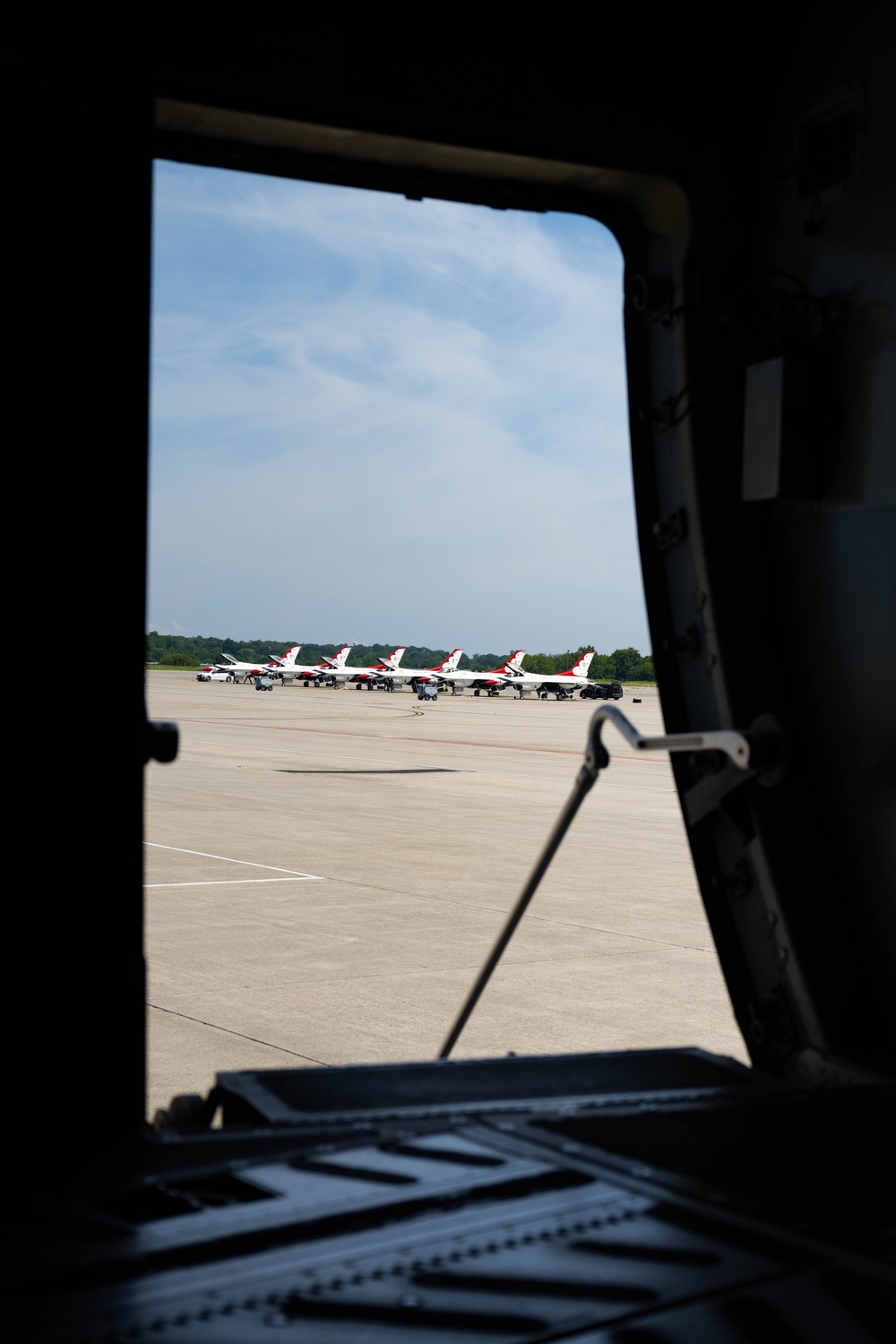 The image is taken from inside a large military aircraft, looking out through an open door or hatch onto a flight line. Visible on the tarmac are multiple aircraft with distinctive red, white, and blue livery consistent with the U.S. Air Force Thunderbirds aerial demonstration team. The aircraft are parked in a row on the apron with ground crew personnel visible nearby.