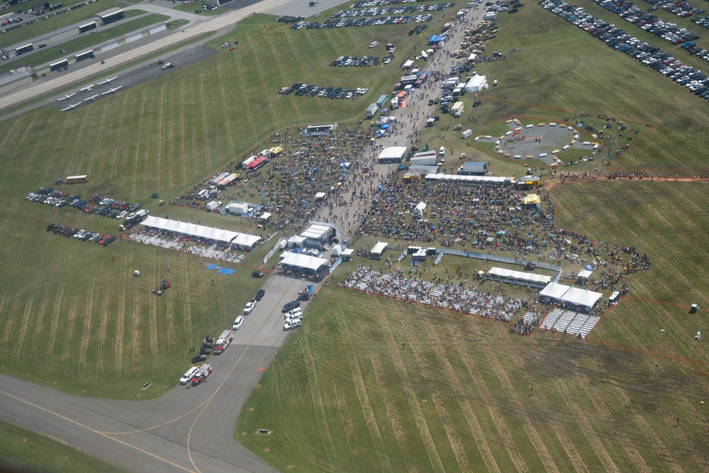 An aerial photograph of a large outdoor event held on the grounds of what appears to be an airfield or military base. The event features multiple white tent structures, large crowds of attendees, rows of seating, vendor areas, and a circular track or display area. Extensive parking lots with numerous vehicles are visible surrounding the event grounds.