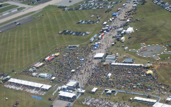 An aerial photograph of a large outdoor event held on the grounds of what appears to be an airfield or military base. The event features multiple white tent structures, large crowds of attendees, rows of seating, vendor areas, and a circular track or display area. Extensive parking lots with numerous vehicles are visible surrounding the event grounds.
