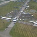 An aerial photograph of a large outdoor event held on the grounds of what appears to be an airfield or military base. The event features multiple white tent structures, large crowds of attendees, rows of seating, vendor areas, and a circular track or display area. Extensive parking lots with numerous vehicles are visible surrounding the event grounds.