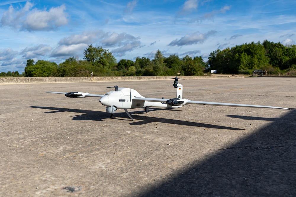 A white fixed-wing UAV (unmanned aerial vehicle) with a pusher propeller configuration is parked on a concrete surface, likely a tarmac or airfield apron. The drone features long glider-style wings, a belly-mounted sensor pod, and what appears to be a German flag marking on the tail. The setting is an open outdoor area with trees and a partly cloudy sky in the background.