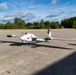 A white fixed-wing UAV (unmanned aerial vehicle) with a pusher propeller configuration is parked on a concrete surface, likely a tarmac or airfield apron. The drone features long glider-style wings, a belly-mounted sensor pod, and what appears to be a German flag marking on the tail. The setting is an open outdoor area with trees and a partly cloudy sky in the background.