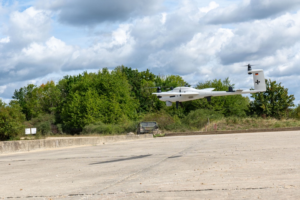 A white unmanned aerial vehicle (UAV/drone) is captured in low-altitude flight over a concrete apron or runway area. The aircraft displays a German Bundeswehr Iron Cross insignia and a small German flag on its tail fin, indicating military affiliation. A military vehicle is visible in the background near a tree line.