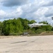 A white unmanned aerial vehicle (UAV/drone) is captured in low-altitude flight over a concrete apron or runway area. The aircraft displays a German Bundeswehr Iron Cross insignia and a small German flag on its tail fin, indicating military affiliation. A military vehicle is visible in the background near a tree line.