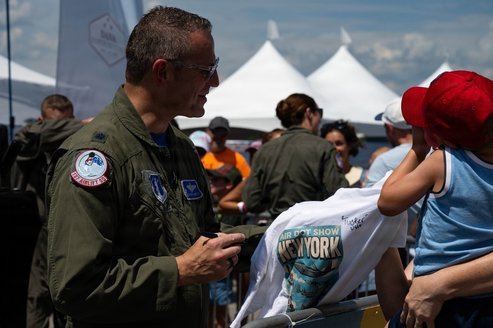 A military aviator in an olive green flight suit is signing autographs at what appears to be the New York Air Show. He is signing a white t-shirt that reads 'Air Dot Show New York' being held by a child wearing a red baseball cap. The event is taking place outdoors with white event tents visible in the background and a crowd of attendees.